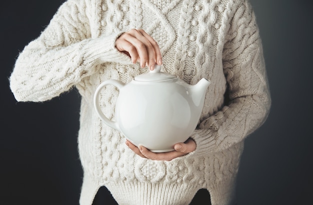 Mujer en suéter caliente sostiene una gran tetera blanca con té en forma de corazón. Vista frontal, mesa de madera grunge. anfas, sin rostro.