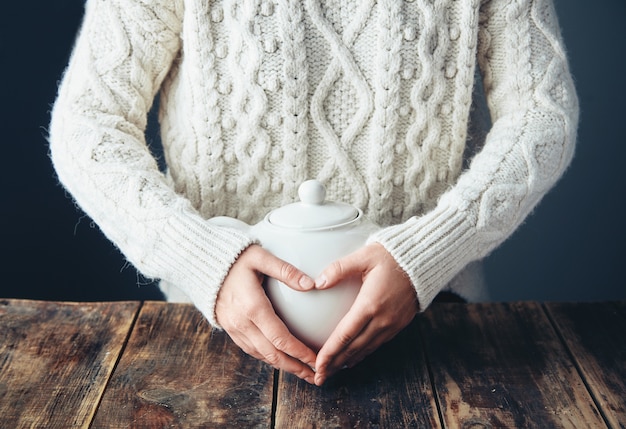 Mujer en suéter caliente tiene las manos en la gran tetera blanca con té en forma de corazón. Vista frontal, mesa de madera grunge. anfas, sin rostro.