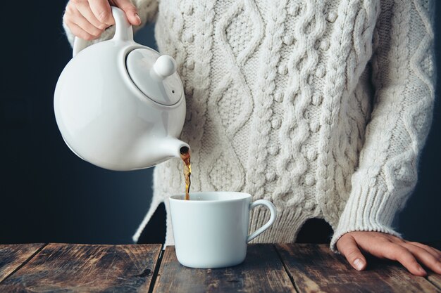 Mujer en suéter grueso de punto cálido vierte té negro de la tetera blanca grande a la taza en la mesa de madera de grunge. Vista frontal, anfas, sin rostro.