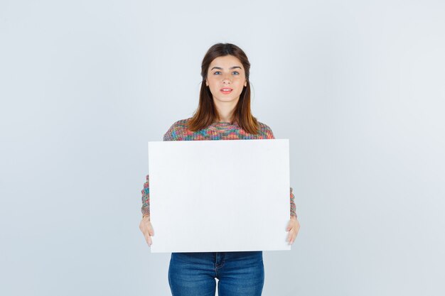 mujer en suéter, jeans manteniendo el tablero en blanco y mirando feliz, vista frontal.