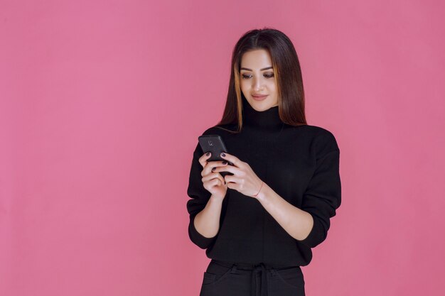 Mujer en suéter negro sosteniendo un teléfono inteligente y enviando mensajes de texto o revisando las redes sociales.