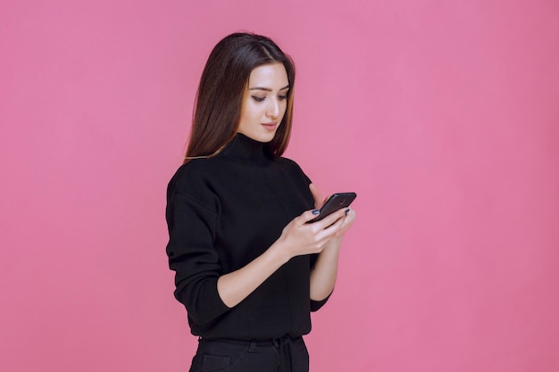 Mujer en suéter negro sosteniendo un teléfono inteligente y enviando mensajes de texto o revisando las redes sociales.