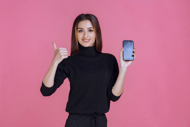 Mujer en suéter negro sosteniendo un teléfono inteligente y haciendo pulgar hacia arriba.