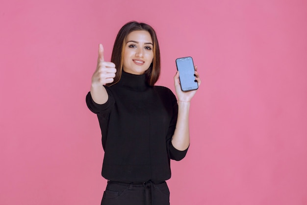 Mujer en suéter negro sosteniendo un teléfono inteligente y haciendo pulgar hacia arriba.