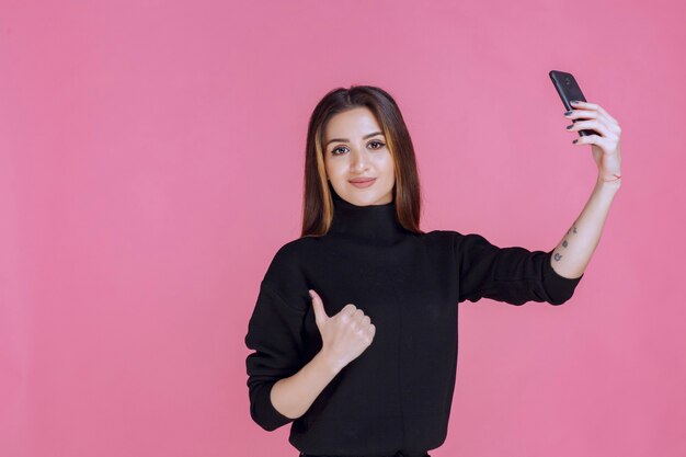 Mujer en suéter negro sosteniendo un teléfono inteligente y tomando su selfie.