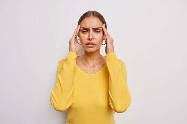 mujer sufre de dolor de cabeza mantiene las manos en las sienes frustrada por el fracaso muecas de dolor doloroso necesita analgésicos usa un jersey amarillo casual sobre blanco