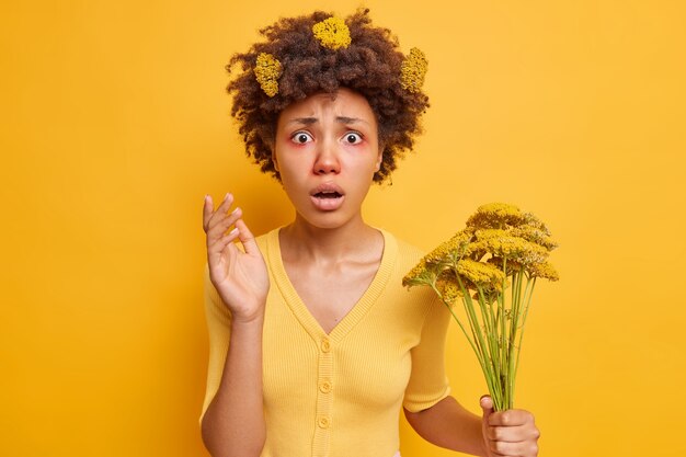 La mujer sufre de fiebre del heno se ve conmocionada tiene una reacción alérgica en las flores silvestres tiene ojos rojos con picazón usa un jersey casual aislado sobre amarillo