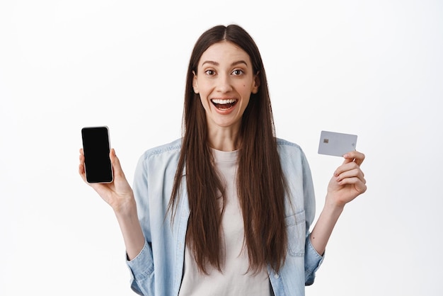 Mujer súper feliz mostrando la pantalla de su teléfono inteligente vacía y la tarjeta de crédito pagando compras en línea en una aplicación de pie sobre fondo blanco