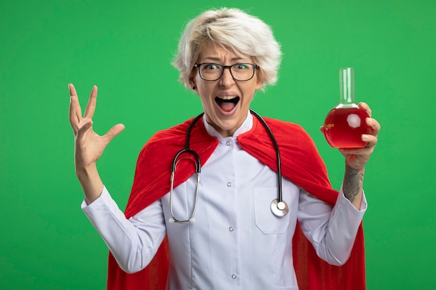 Mujer de superhéroe eslava ansiosa en uniforme médico con capa roja y estetoscopio en gafas ópticas se encuentra con la mano levantada y sostiene líquido químico rojo en frasco de vidrio en pared verde