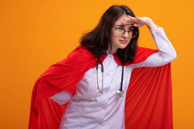 Mujer de superhéroe joven confiada con uniforme médico y un estetoscopio con gafas manteniendo la mano detrás de la espalda y en la frente mirando al lado en la distancia aislada en la pared