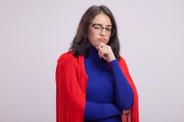 Mujer de superhéroe joven pensativa en capa roja con gafas manteniendo la mano en la barbilla mirando hacia abajo aislado en la pared blanca con espacio de copia