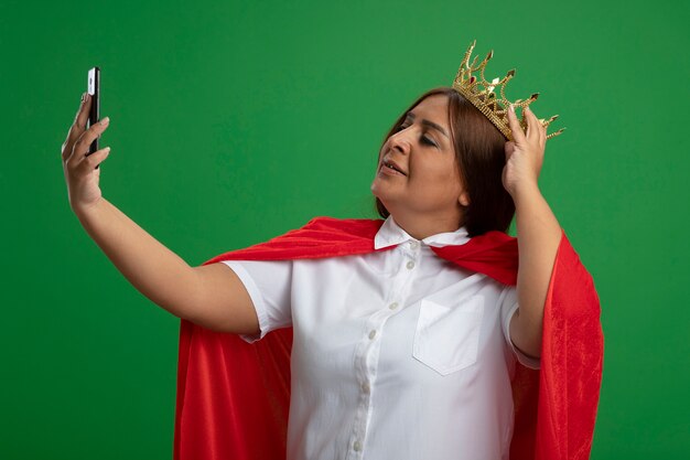 Foto gratuita mujer superhéroe de mediana edad complacida con corona tomar un selfie poniendo la mano en la corona aislada en verde