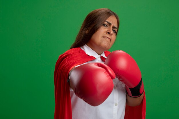 Mujer de superhéroe de mediana edad confiada con guantes de boxeo sosteniendo la mano aislada en verde