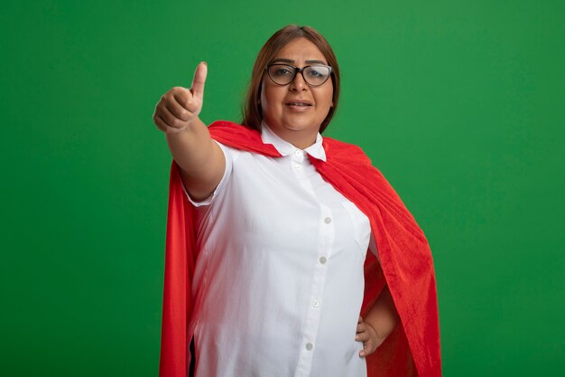 Mujer de superhéroe de mediana edad disgustada con gafas mostrando el pulgar hacia arriba aislado sobre fondo verde