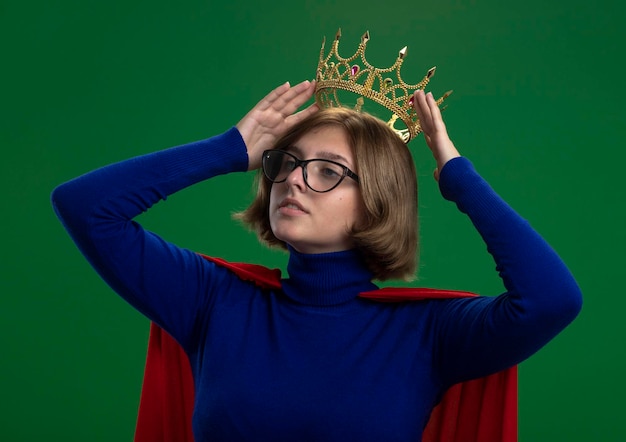 Foto gratuita mujer de superhéroe rubia joven confiada en capa roja con gafas mirando al lado sosteniendo la corona sobre la cabeza aislada en la pared verde