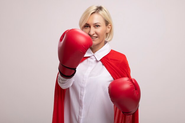 Mujer de superhéroe rubia de mediana edad alegre en capa roja con guantes de boxeo manteniendo los puños en el aire