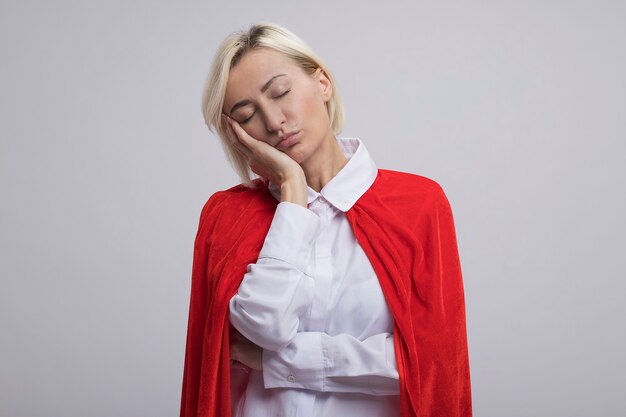 Mujer de superhéroe rubia de mediana edad cansada en capa roja poniendo la mano en la cara con los ojos cerrados aislado en la pared blanca con espacio de copia