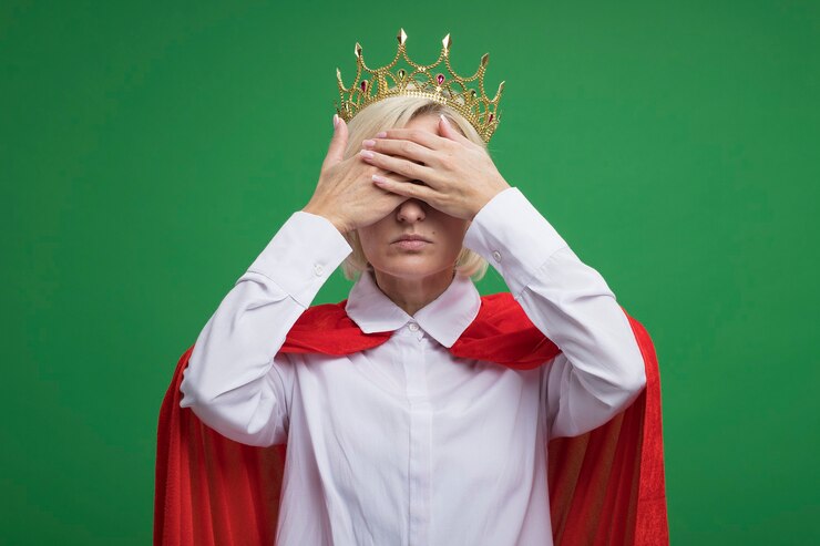 Foto gratuito mujer superhéroe rubia de mediana edad en capa roja con gafas y corona cubriendo los ojos con las manos