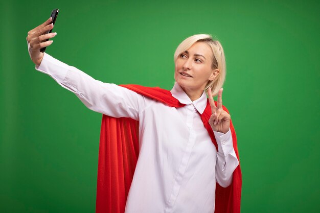 Mujer superhéroe rubia de mediana edad complacida en capa roja haciendo el signo de la paz tomando selfie aislado en la pared verde con espacio de copia