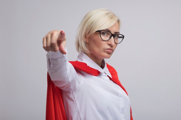 Mujer de superhéroe rubia de mediana edad confiada en capa roja con gafas de pie en la vista de perfil mirando y apuntando aislado en la pared blanca con espacio de copia