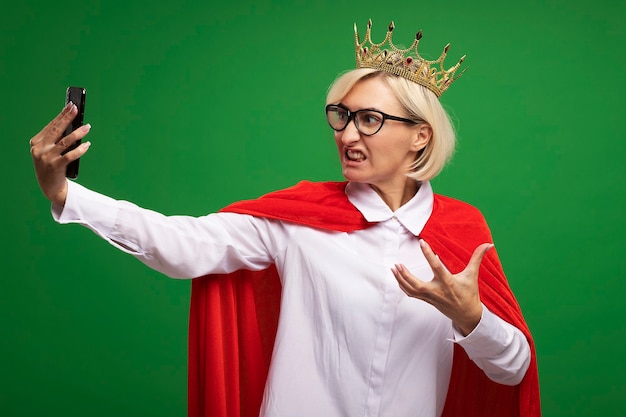 Mujer de superhéroe rubia de mediana edad molesta en capa roja con gafas y corona manteniendo la mano en el aire tomando selfie aislado en la pared verde