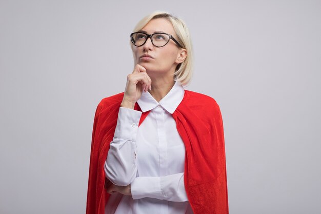 Mujer de superhéroe rubia de mediana edad pensativa en capa roja con gafas poniendo la mano en la barbilla mirando hacia arriba