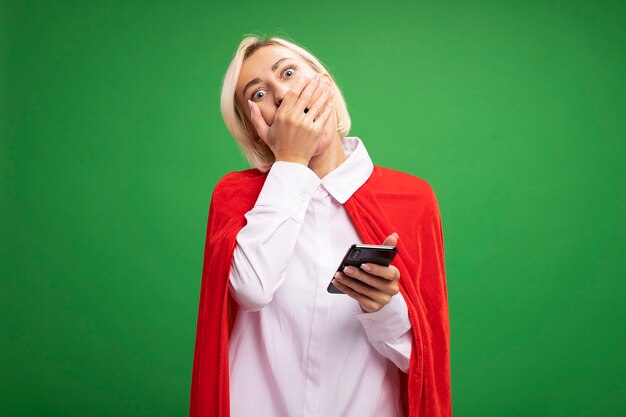 Mujer de superhéroe rubia de mediana edad preocupada en capa roja sosteniendo el teléfono móvil manteniendo la mano en la boca aislada en la pared verde con espacio de copia