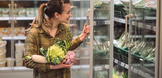 Mujer en el supermercado Hermosa joven tiene en las manos vegetales orgánicos frescos y abre la nevera en el supermercado