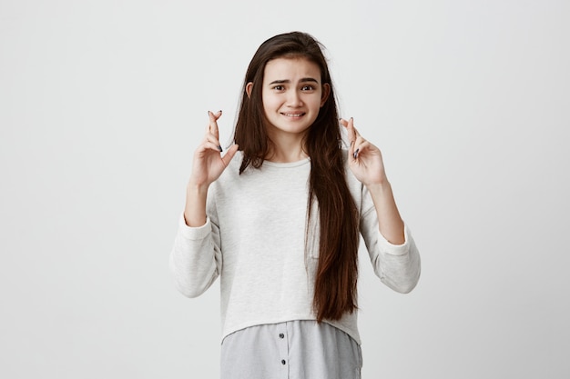 Mujer supersticiosa con cabello oscuro y liso y cara bonita, apretando los dientes, cruzando los dedos para tener buena suerte, esperando que sus deseos se hagan realidad, con una mirada excitada. Emociones y sentimientos humanos