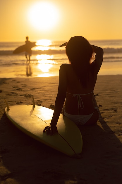 Mujer con tabla de surf protegiéndose los ojos mientras se relaja en la playa