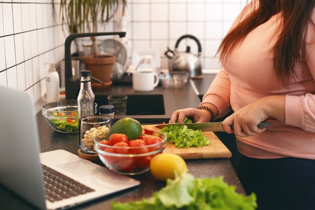 Mujer de talla grande irreconocible de pie en el mostrador de la cocina y cortando verduras orgánicas frescas en una tabla de cortar, haciendo una ensalada saludable y baja en calorías mientras ve una película en línea en una computadora portátil