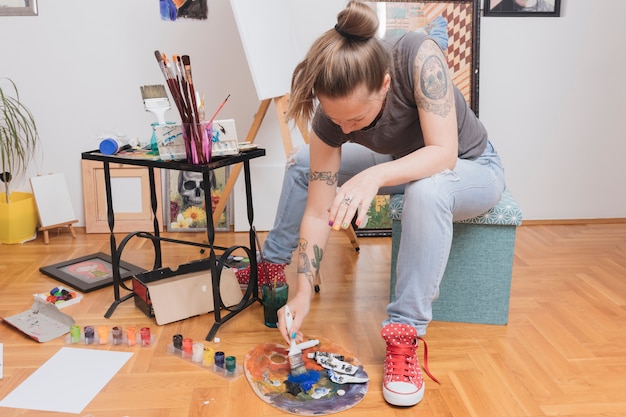 Foto gratuita mujer tatuada joven que se sienta en colores de mezcla del taburete en la paleta