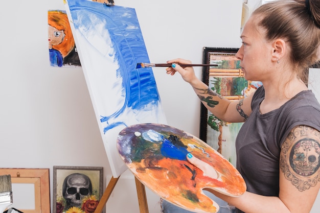 Mujer tatuada de pie y pintando cuadro azul sobre lienzo.