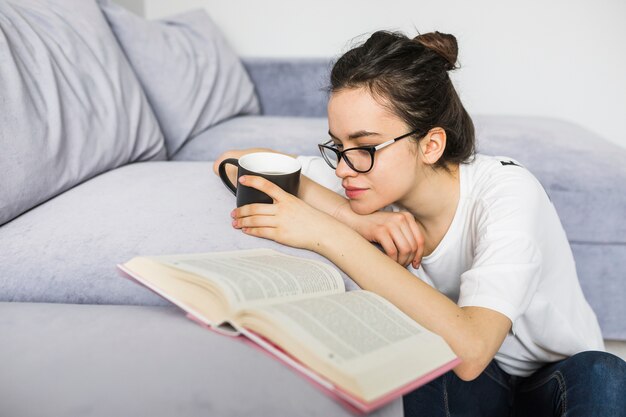 Mujer con taza apoyándose en el sofá cerca del libro