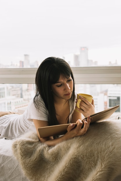 Foto gratuita mujer con taza de lectura junto a la ventana