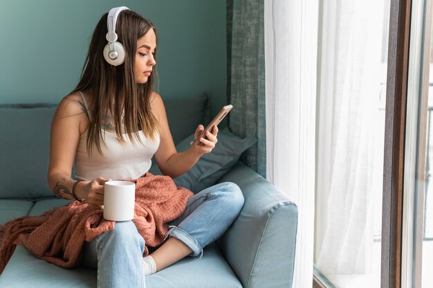 Mujer con teléfono inteligente con auriculares en casa mientras toma café durante la pandemia