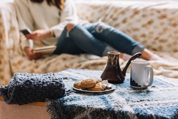 Mujer con teléfono inteligente en el sofá junto a la mesa con bebida y galletas