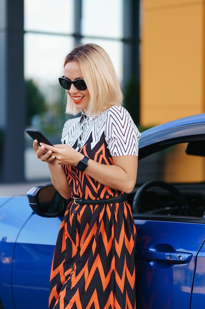 Mujer con teléfono móvil, comunicación o aplicación en línea, de pie cerca del automóvil en la calle de la ciudad o estacionamiento, al aire libre. Coche compartido, servicio de alquiler