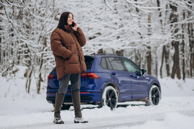 Mujer con teléfono de pie junto a su coche en el bosque de invierno