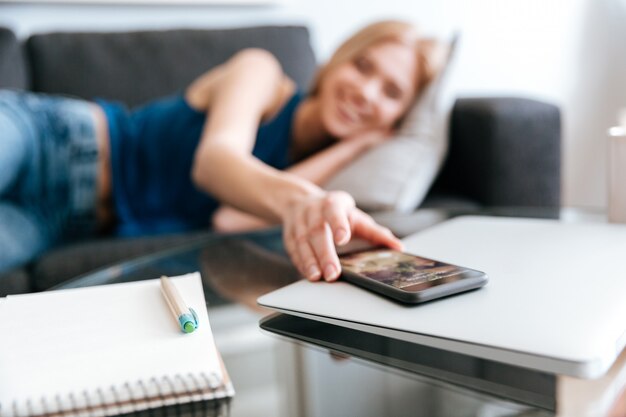 Mujer tendida en el sofá y tomando el teléfono móvil de la mesa