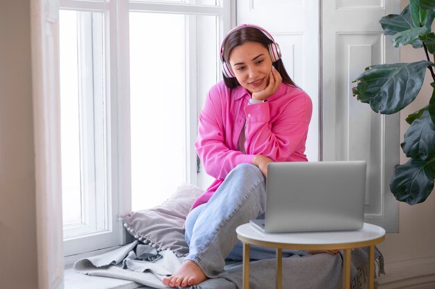Mujer teniendo una videollamada en una computadora portátil mientras está sentada en el alféizar de la ventana en casa