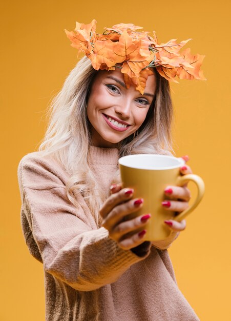 Mujer con tiara de hojas de arce que muestra una taza de café de pie contra una pared amarilla