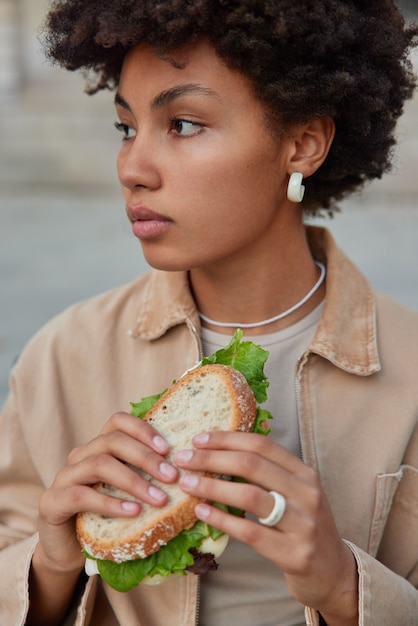 Mujer tiene bocadillos en la calle tiene delicioso sándwich come sabrosa comida rápida mira a un lado pensativamente vestida con ropa de moda plantea fuera