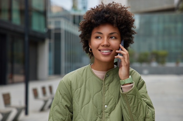 La mujer tiene conversación telefónica se encuentra afuera en el entorno urbano satisfecho con las tarifas en roaming viste chaqueta