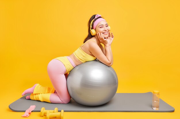 mujer tiene entrenamiento físico con pelota suiza sonríe gratamente vestida con ropa deportiva escucha música a través de auriculares usa equipo deportivo