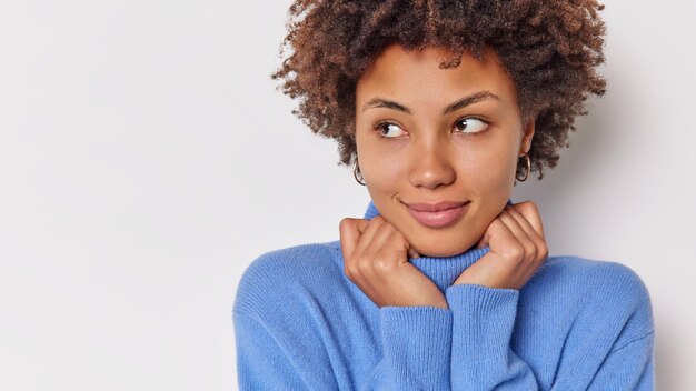 La mujer tiene una expresión de ensueño, mantiene las manos en el cuello del suéter azul y está sumido en pensamientos aislados en blanco con un espacio en blanco para publicidad.