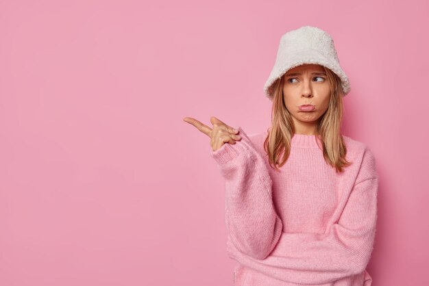 La mujer tiene una expresión de mal humor frunce los labios y señala con el dedo índice viste un sombrero de piel blanco y un jersey aislado en rosa