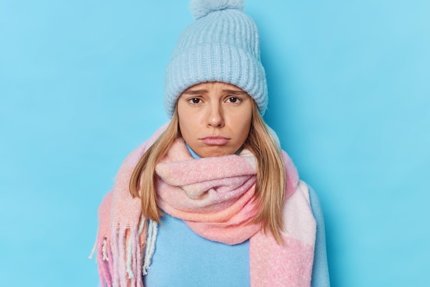 Foto gratuita la mujer tiene una expresión sombría cuando alguien lastimó sus sentimientos se queja de que algo usa un gorro de punto y una bufanda alrededor del cuello aislado en azul se siente culpable