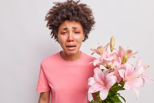 La mujer tiene los ojos rojos hinchados reacciona a los alérgenos tiene ramo de lirios viste camiseta plantea interior