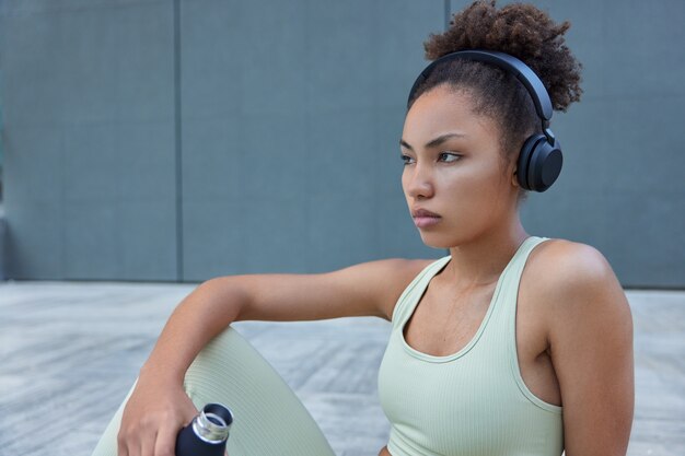 Mujer tiene sudor después de un entrenamiento cardiovascular intensivo toma un descanso bebe agua refrescante de botella vestida con ropa deportiva escucha música motivacional